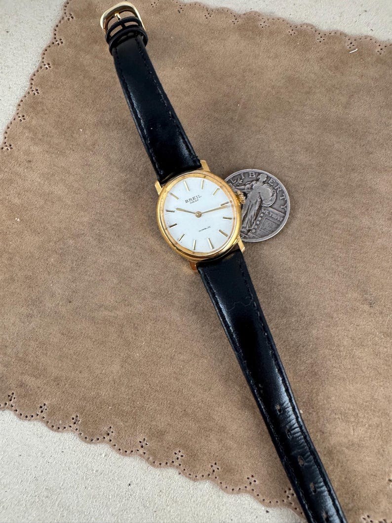 K&ouml;nnte beinhalten: Eine goldfarbene Breil-Uhr mit wei&szlig;em Zifferblatt und schwarzem Lederarmband. Die Uhr liegt auf einer braunen Wildlederoberfl&auml;che.