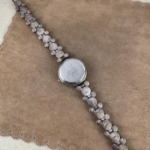 Puede incluir: Reloj plateado con esfera redonda y una correa con forma de Mickey Mouse. La esfera del reloj tiene el texto "JAZ" y "WATER RESISTANT 50M" impreso en ella.