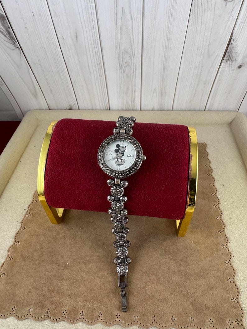 Puede incluir: Un reloj plateado con una esfera blanca y un dise&ntilde;o de Mickey Mouse. El reloj tiene una correa plateada con un dise&ntilde;o de Mickey Mouse.