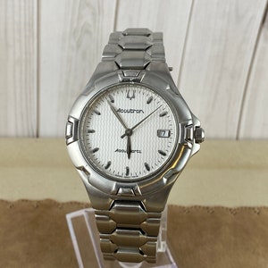 Op de afbeelding: Zilveren Accutron quartz polshorloge met een witte, getextureerde wijzerplaat en een datumvenster. De band is van roestvrij staal. De wijzerplaat toont 'Accutron'. Het horloge wordt op een transparante standaard gepresenteerd.