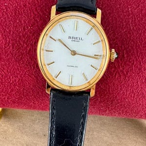 K&ouml;nnte beinhalten: Eine goldfarbene Breil Okay Armbanduhr mit wei&szlig;em Zifferblatt und schwarzem Lederarmband. Das Zifferblatt hat schwarze r&ouml;mische Ziffern als Stundenmarkierungen und schwarze Zeiger. Auf dem Zifferblatt sind au&szlig;erdem die Texte "Breil Okay" und "Incabloc" aufgedruckt.