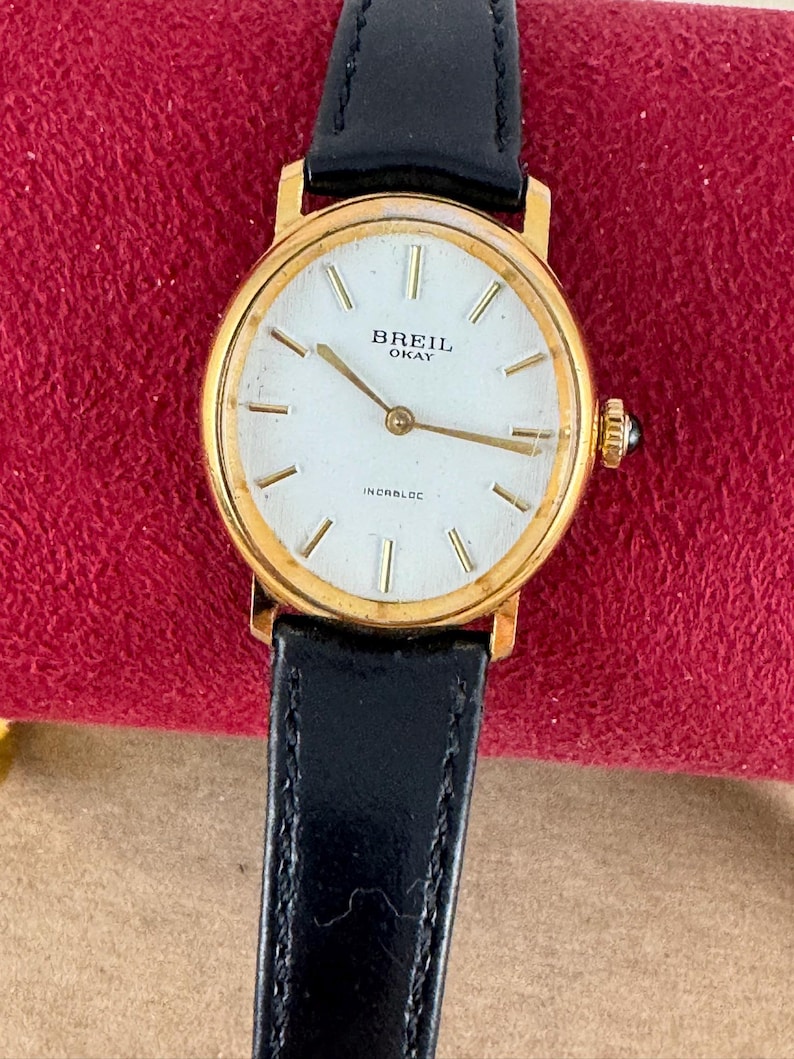 K&ouml;nnte beinhalten: Eine goldfarbene Breil Okay Armbanduhr mit wei&szlig;em Zifferblatt und schwarzem Lederarmband. Das Zifferblatt hat schwarze r&ouml;mische Ziffern als Stundenmarkierungen und schwarze Zeiger. Auf dem Zifferblatt sind au&szlig;erdem die Texte "Breil Okay" und "Incabloc" aufgedruckt.