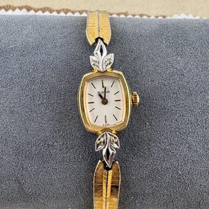 Puede incluir: Un reloj de pulsera Bulova vintage con caja y correa doradas. La esfera es rectangular con fondo blanco y agujas negras. La correa presenta detalles decorativos en forma de hojas con pequeños adornos con forma de diamante.