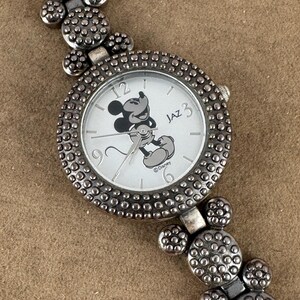 Puede incluir: Reloj de Mickey Mouse plateado con una esfera de Mickey Mouse en blanco y negro y una correa plateada con orejas de Mickey Mouse. La esfera del reloj dice "Jaz" y "Disney".
