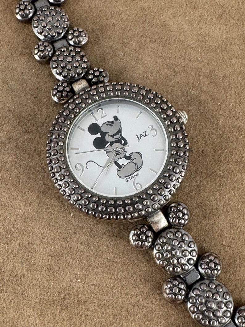 Puede incluir: Reloj de Mickey Mouse plateado con una esfera de Mickey Mouse en blanco y negro y una correa plateada con orejas de Mickey Mouse. La esfera del reloj dice "Jaz" y "Disney".