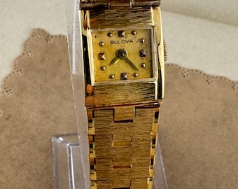 May include: A vintage Bulova watch with a gold-tone rectangular face and band. The watch face has a textured gold background with black hands and the Bulova brand name. The watch band is also gold-toned and has a textured design.