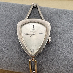 May include: A vintage Bucherer wristwatch with a silver-toned triangular face and a silver band. The watch face has a white background with black hands and markers. The watch is marked "Swiss Made".