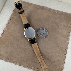 K&ouml;nnte beinhalten: Eine goldfarbene Armbanduhr mit einem schwarzen Lederarmband. Das Zifferblatt der Uhr ist rund und silberfarben. Das Armband tr&auml;gt den Schriftzug "Swiss Made". Die Uhr liegt auf einer braunen Oberfl&auml;che.