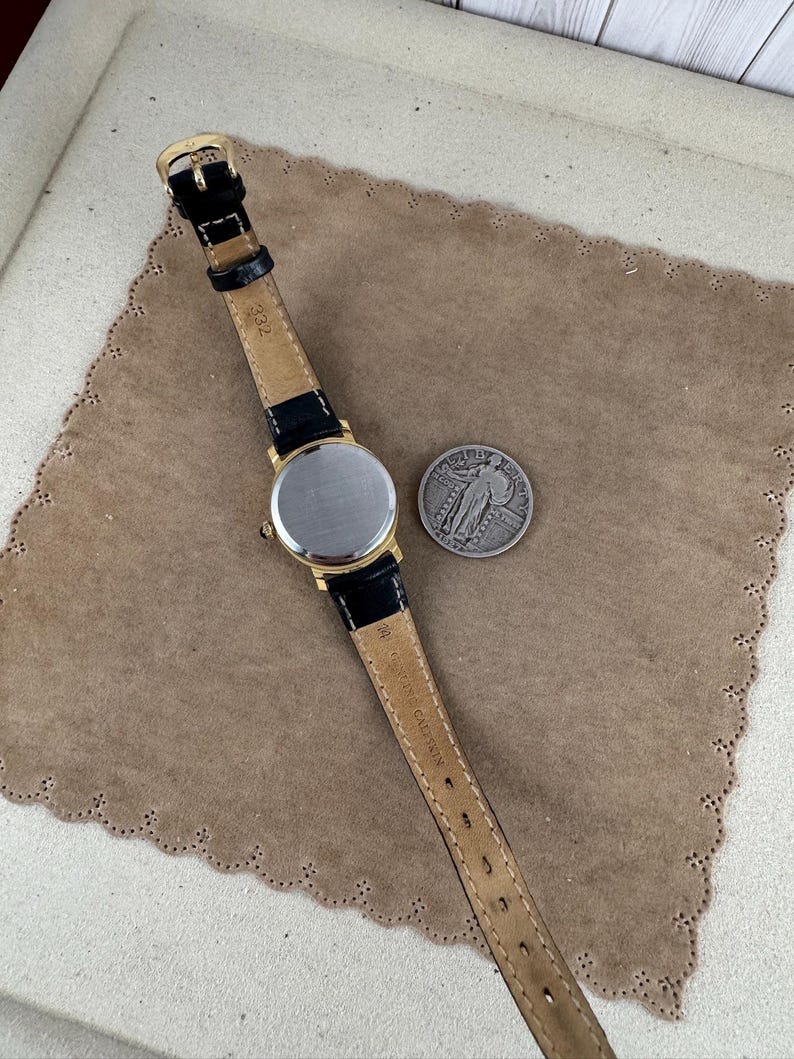 K&ouml;nnte beinhalten: Eine goldfarbene Armbanduhr mit einem schwarzen Lederarmband. Das Zifferblatt der Uhr ist rund und silberfarben. Das Armband tr&auml;gt den Schriftzug "Swiss Made". Die Uhr liegt auf einer braunen Oberfl&auml;che.