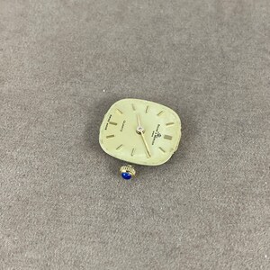 May include: A gold-toned square watch face with black hands and Roman numeral markings. The watch face reads "QUARTZ" and "SWISS MADE" and is labeled "Eterna Matic".