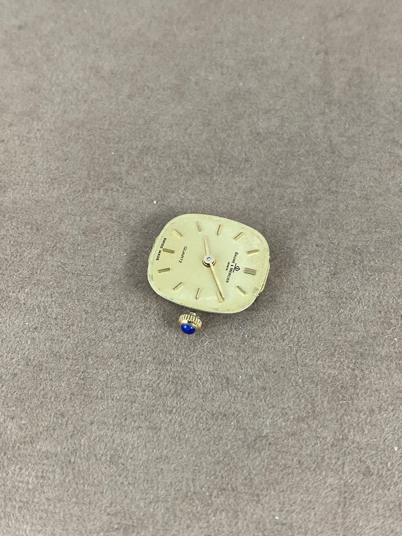 May include: A gold-toned square watch face with black hands and Roman numeral markings. The watch face reads "QUARTZ" and "SWISS MADE" and is labeled "Eterna Matic".