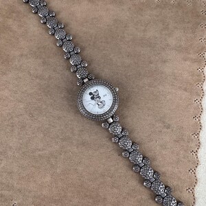 Puede incluir: Un reloj plateado con una cara de Mickey Mouse en la esfera. La correa del reloj est&aacute; hecha de cabezas de Mickey Mouse enlazadas con diamantes de imitaci&oacute;n.