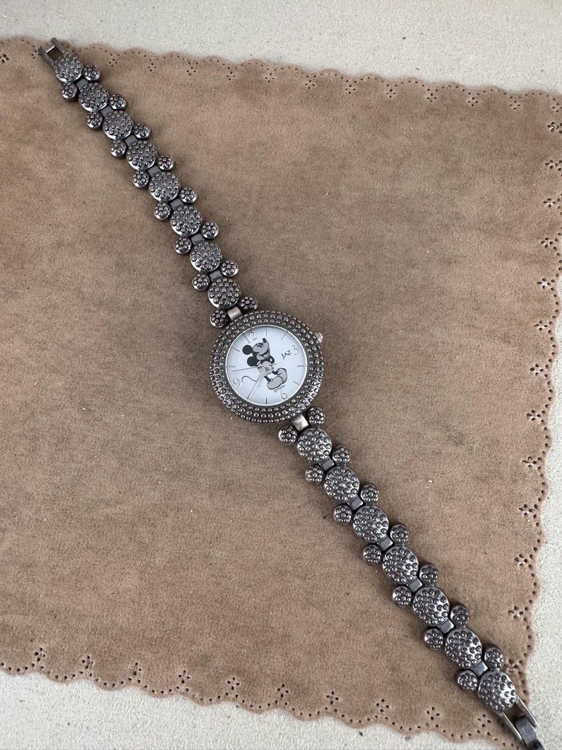 Puede incluir: Un reloj plateado con una cara de Mickey Mouse en la esfera. La correa del reloj est&aacute; hecha de cabezas de Mickey Mouse enlazadas con diamantes de imitaci&oacute;n.