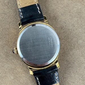 K&ouml;nnte beinhalten: Eine goldfarbene Armbanduhr mit einem schwarzen Lederarmband. Das Zifferblatt der Uhr ist rund und hat eine silberfarbene Oberfl&auml;che. Auf der R&uuml;ckseite der Uhr sind die Worte "Eberhard & Co.  acier" eingraviert.