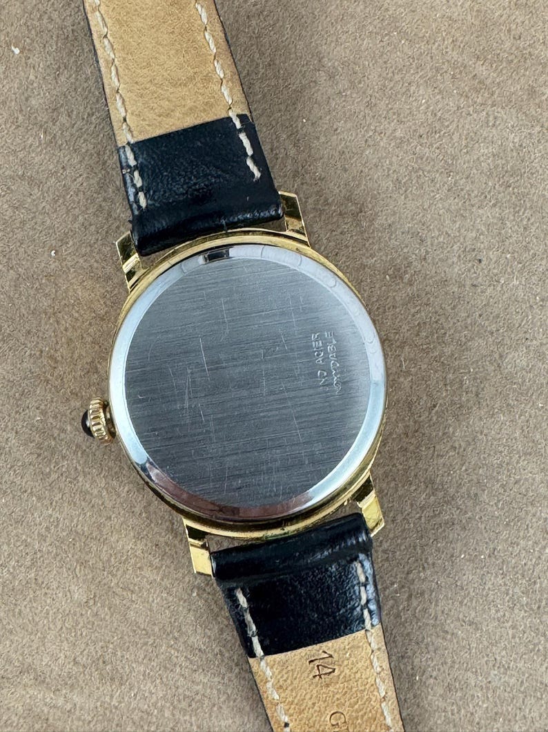 K&ouml;nnte beinhalten: Eine goldfarbene Armbanduhr mit einem schwarzen Lederarmband. Das Zifferblatt der Uhr ist rund und hat eine silberfarbene Oberfl&auml;che. Auf der R&uuml;ckseite der Uhr sind die Worte "Eberhard & Co.  acier" eingraviert.