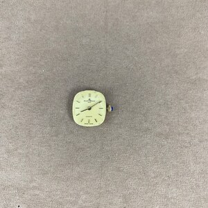 May include: A square-shaped watch face with a gold-colored case and a white dial. The watch face has black Roman numeral hour markers and black hands. The watch face also has the text "Quartz" and "Swiss Made" printed on it.