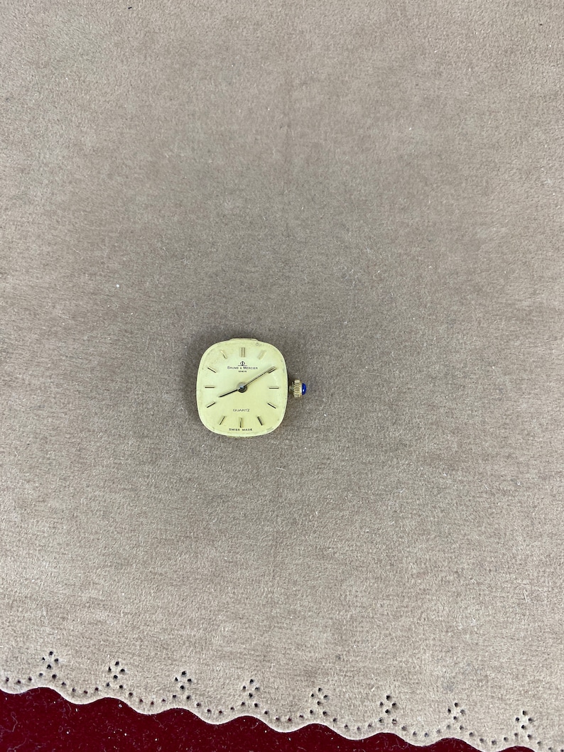 May include: A square-shaped watch face with a gold-colored case and a white dial. The watch face has black Roman numeral hour markers and black hands. The watch face also has the text "Quartz" and "Swiss Made" printed on it.