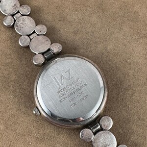 Puede incluir: Un reloj de pulsera plateado con una esfera redonda y una correa plateada. La esfera del reloj tiene la palabra "JAZ" impresa en ella. La parte posterior del reloj tiene el texto "Water Resistant, Base Metal Bezel, Stainless Steel Back, V301-X0822, Mov't Japan".