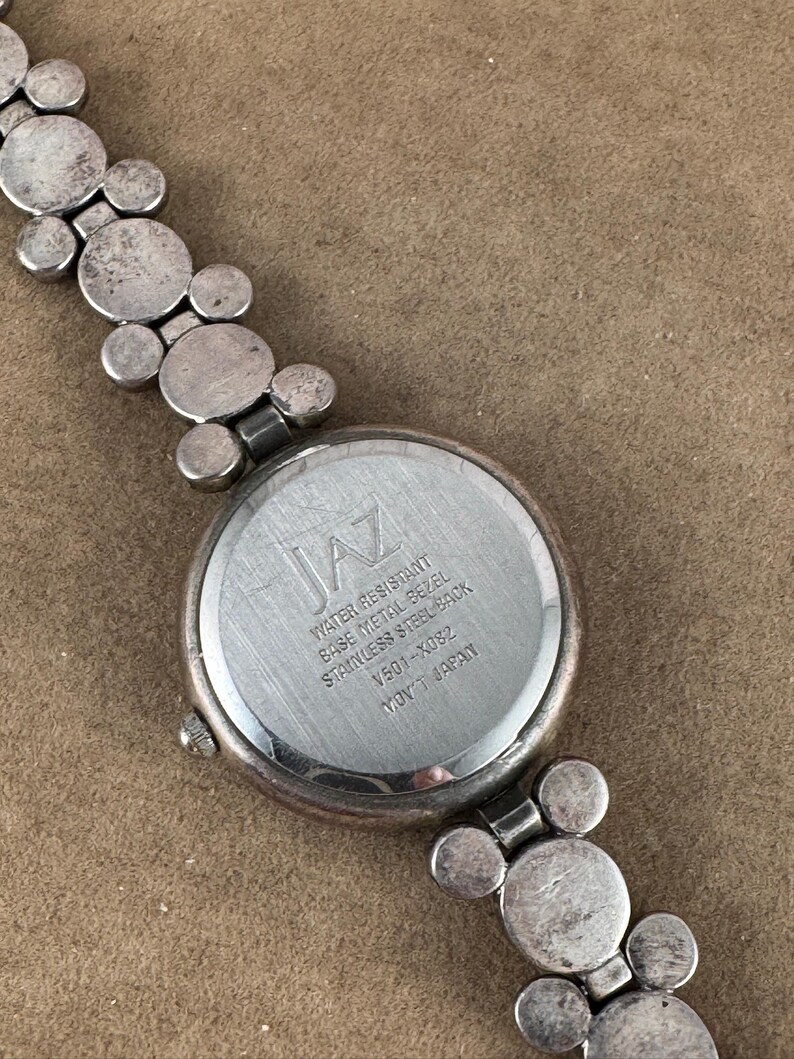 Puede incluir: Un reloj de pulsera plateado con una esfera redonda y una correa plateada. La esfera del reloj tiene la palabra "JAZ" impresa en ella. La parte posterior del reloj tiene el texto "Water Resistant, Base Metal Bezel, Stainless Steel Back, V301-X0822, Mov't Japan".