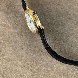 K&ouml;nnte beinhalten: Eine goldfarbene Armbanduhr mit wei&szlig;em Zifferblatt und schwarzem Lederarmband. Die Uhr hat einen schwarzen Stein oben auf dem Zifferblatt.