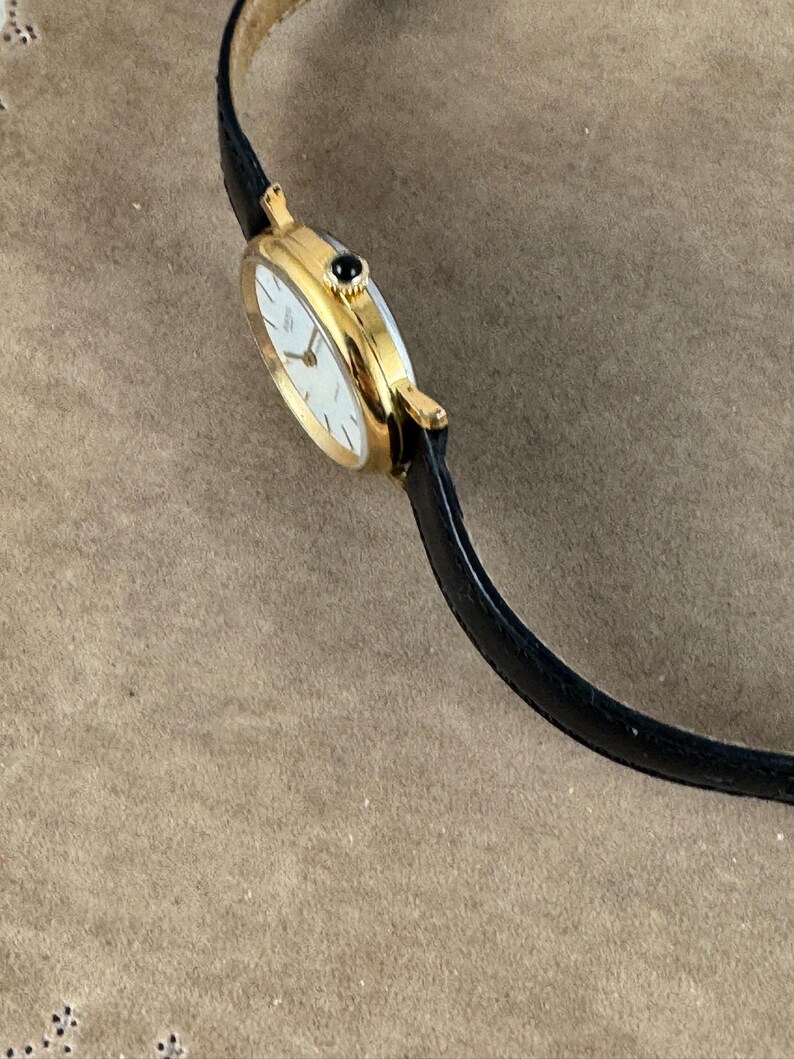 K&ouml;nnte beinhalten: Eine goldfarbene Armbanduhr mit wei&szlig;em Zifferblatt und schwarzem Lederarmband. Die Uhr hat einen schwarzen Stein oben auf dem Zifferblatt.