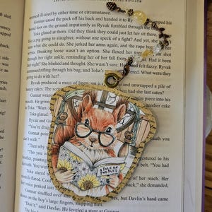 May include: A whimsical bookmark featuring a watercolor illustration of a squirrel wearing glasses and reading a book titled "A Tale of Two Nuts." The bookmark has a beaded charm attached.