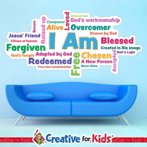 Op de afbeelding: Een blauwe bank met twee kussens staat onder een muursticker. De sticker toont de woorden "I Am" in grote blauwe letters, omringd door woorden als "Forgiven", "Blessed" en "Chosen". De sticker is veelkleurig.