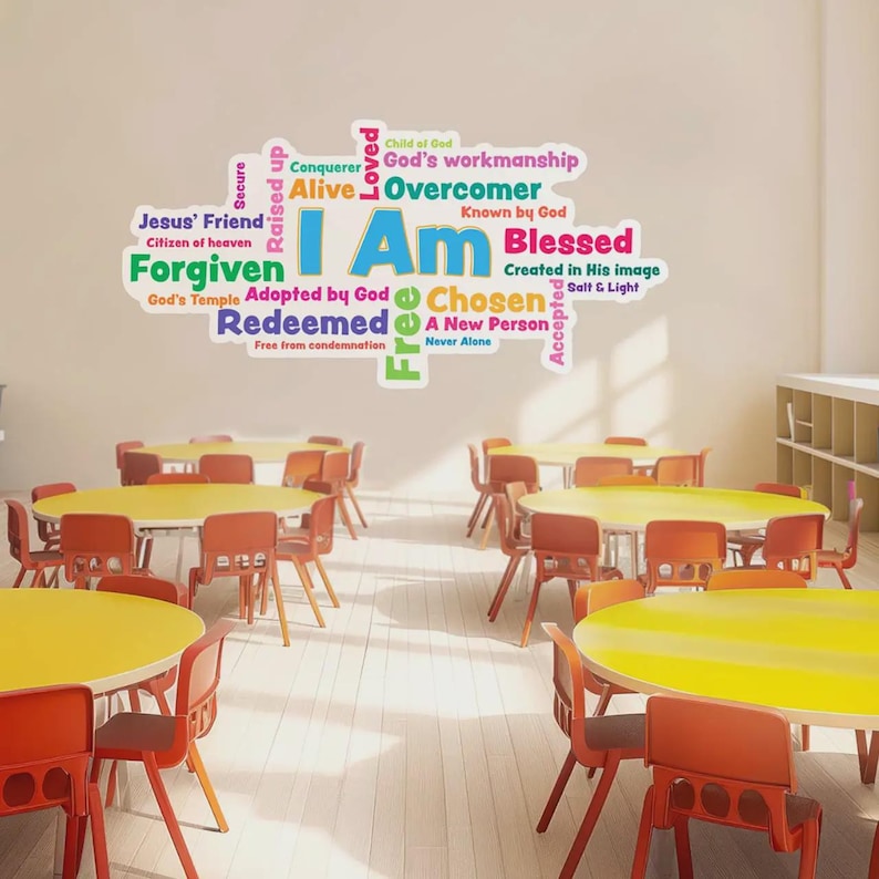 Op de afbeelding: Een klaslokaal met ronde tafels met gele bladen en oranje stoelen. Een muursticker toont de woorden "I Am" in grote blauwe letters, omringd door inspirerende woorden als "Forgiven" en "Blessed".