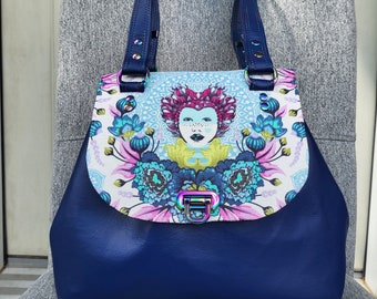 May include: A blue leather tote bag with a floral print and a colorful face design. The bag has a flap closure with a metal clasp.