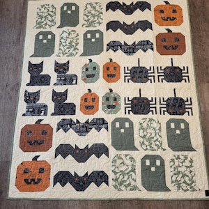 May include: A rectangular quilt featuring a cream-coloured background with a repeating pattern of Halloween-themed fabric blocks. The design includes dark grey bats, orange jack-o'-lanterns, black cats, dark grey spiders, and green or dark grey ghosts.