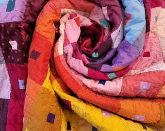 May include: A colorful patchwork quilt with a variety of squares in shades of red, orange, yellow, blue, purple, and pink. The quilt is folded in a spiral shape, creating a unique and eye-catching design.