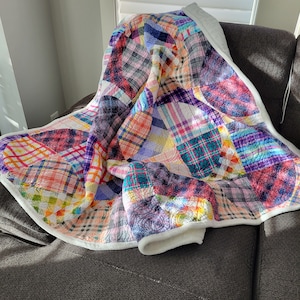 May include: A colourful patchwork quilt with a white border, folded and draped over a brown armchair. The quilt is made of various plaid fabrics in shades of pink, purple, blue, and green.