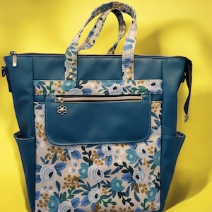 May include: A teal blue tote bag with a floral pattern. The bag has a zipper closure and a top handle. The bag also has a zippered pocket on the front.