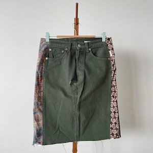 May include: Olive green denim skirt with patterned fabric panels on the sides. The skirt hangs on a wooden hanger. The decorative panels feature floral and geometric patterns in brown, blue, and pink. The skirt is approximately 60 cm long.