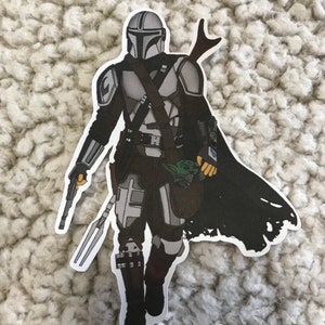 May include: A black and white sticker of a Mandalorian character from the Star Wars franchise. The character is wearing a helmet and armor and is holding a weapon. The character is also holding a small, green creature in a bag.