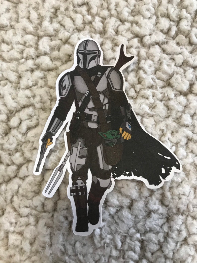 May include: A black and white sticker of a Mandalorian character from the Star Wars franchise. The character is wearing a helmet and armor and is holding a weapon. The character is also holding a small, green creature in a bag.