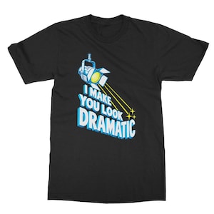 May include: A black short-sleeve t-shirt featuring a graphic of a blue stage spotlight shining a yellow beam of light. Below the light, the text reads I MAKE YOU LOOK DRAMATIC in bold, blue, three-dimensional lettering with white highlights.