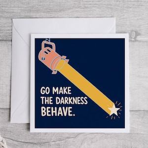 Puede incluir: Una tarjeta de felicitación cuadrada con fondo azul oscuro que muestra un foco color melocotón proyectando un haz de luz amarillo sobre una estrella blanca. El texto de la tarjeta dice: "GO MAKE THE DARKNESS BEHAVE." Un sobre blanco está detrás.
