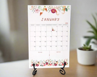 May include: A floral-themed desk calendar for January 2026, displayed on a black metal stand. The calendar features a white background with a grid layout, adorned with watercolor floral designs in pink, red, and green.