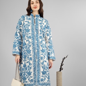 May include: A white quilted coat with a blue floral pattern and a button closure. The coat is long and has a mandarin collar. The model is wearing blue jeans and beige heels.