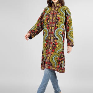May include: A long black embroidered coat with a mandarin collar and floral patterns in shades of red, yellow, green, and blue. The coat is worn over blue jeans.