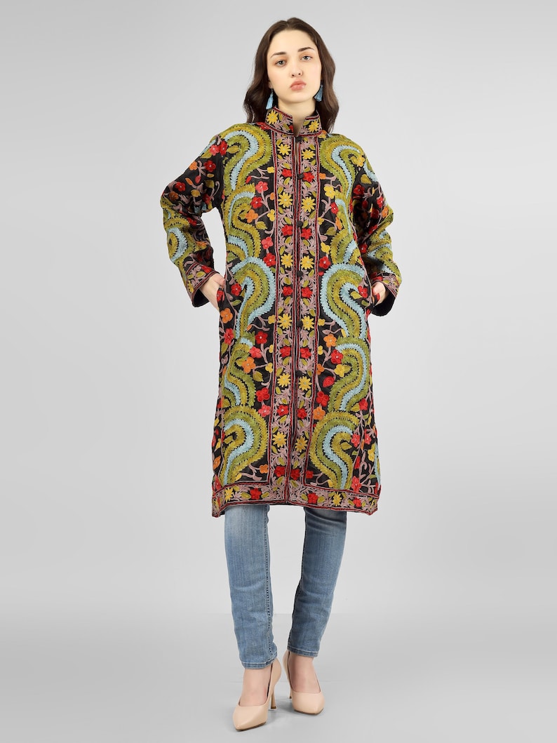 May include: A long black coat with colorful floral embroidery. The coat has a mandarin collar and a button closure. The model is wearing blue jeans and heels.