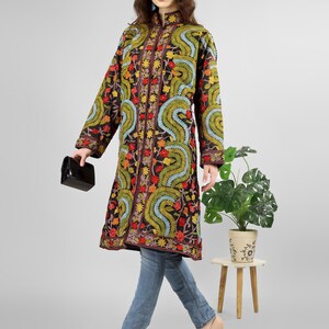 May include: A long black embroidered coat with a floral and paisley design. The coat has a mandarin collar and a button closure. The model is wearing blue jeans and brown heels.