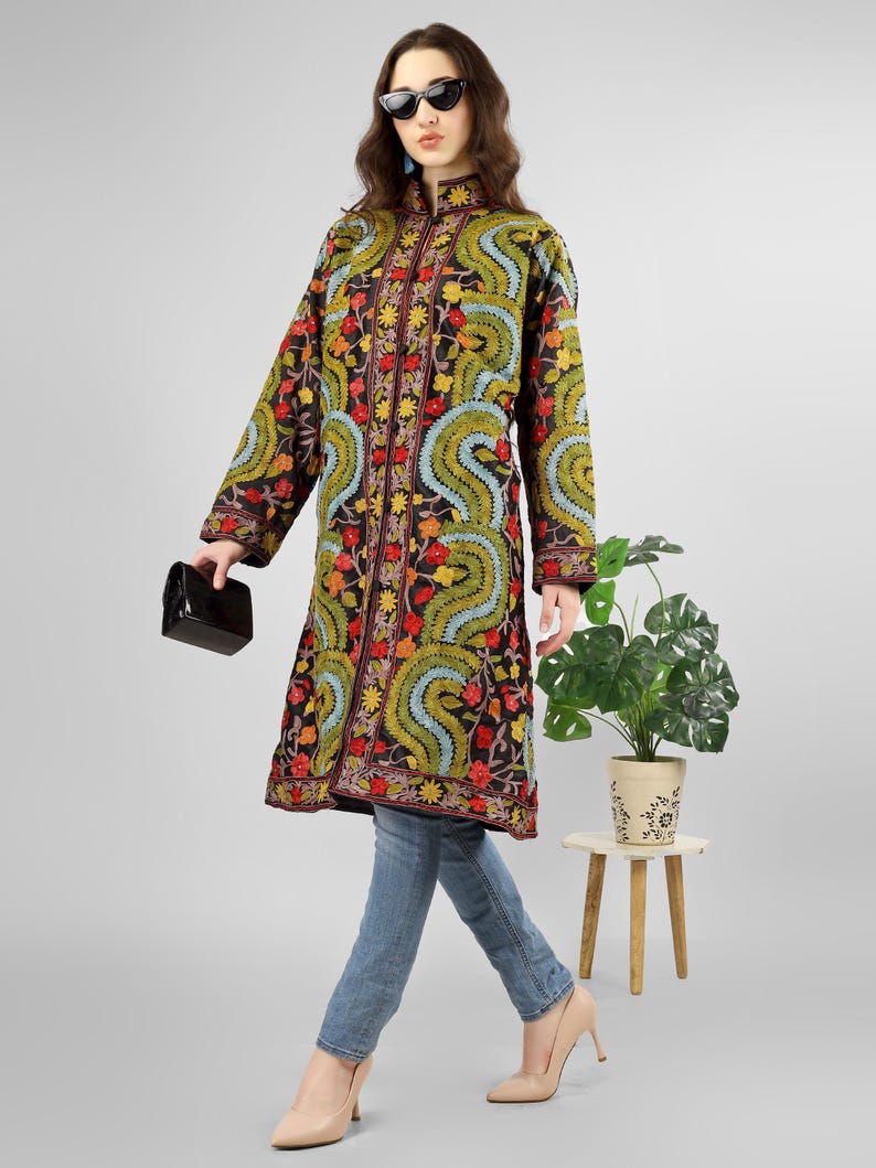 May include: A long black embroidered coat with a floral and paisley design. The coat has a mandarin collar and a button closure. The model is wearing blue jeans and brown heels.