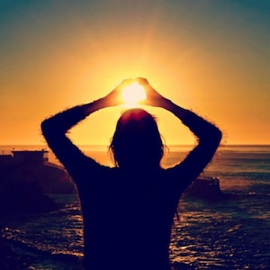 May include: Silhouette of a person framing the setting sun with their hands, creating a heart shape. The person is standing on a rocky shore with the ocean in the background.