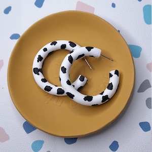 May include: A pair of white hoop earrings with a black and white cow print design. The earrings are on a yellow dish.