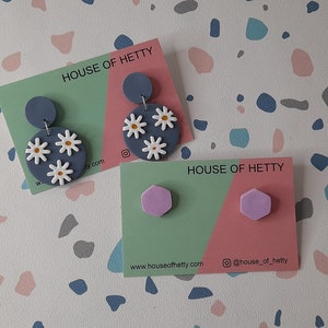 May include: Two pairs of handmade polymer clay earrings. The first pair features a blue circle with white daisy flowers. The second pair features a pair of lavender hexagonal earrings. Both pairs are displayed on cards with the text "House of Hetty" and website information.