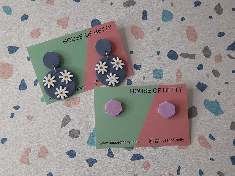 May include: Two pairs of handmade polymer clay earrings. The first pair features a blue circle with white daisy flowers. The second pair features a pair of lavender hexagonal earrings. Both pairs are displayed on cards with the text "House of Hetty" and website information.
