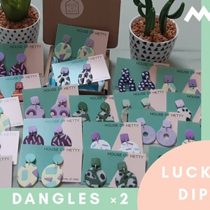 May include: A collection of colorful, geometric patterned polymer clay earrings on display cards. The cards are labeled "House of Hetty" and the earrings are arranged in a random assortment. The text "Lucky Dip" and "Dangles x 2" are visible in the image.