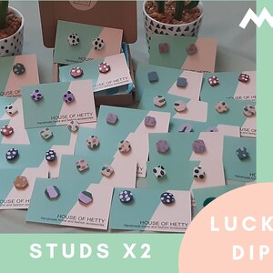 May include: A collection of colorful, patterned polymer clay earrings on display. The earrings are arranged on cards with the text "House of Hetty" and "Lucky Dip" on them. The earrings are available in a variety of colors and patterns, including polka dots, stripes, and geometric shapes.